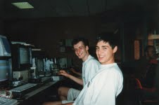 Two teenagers at a desk of computer equipment, turning to smile at the camera